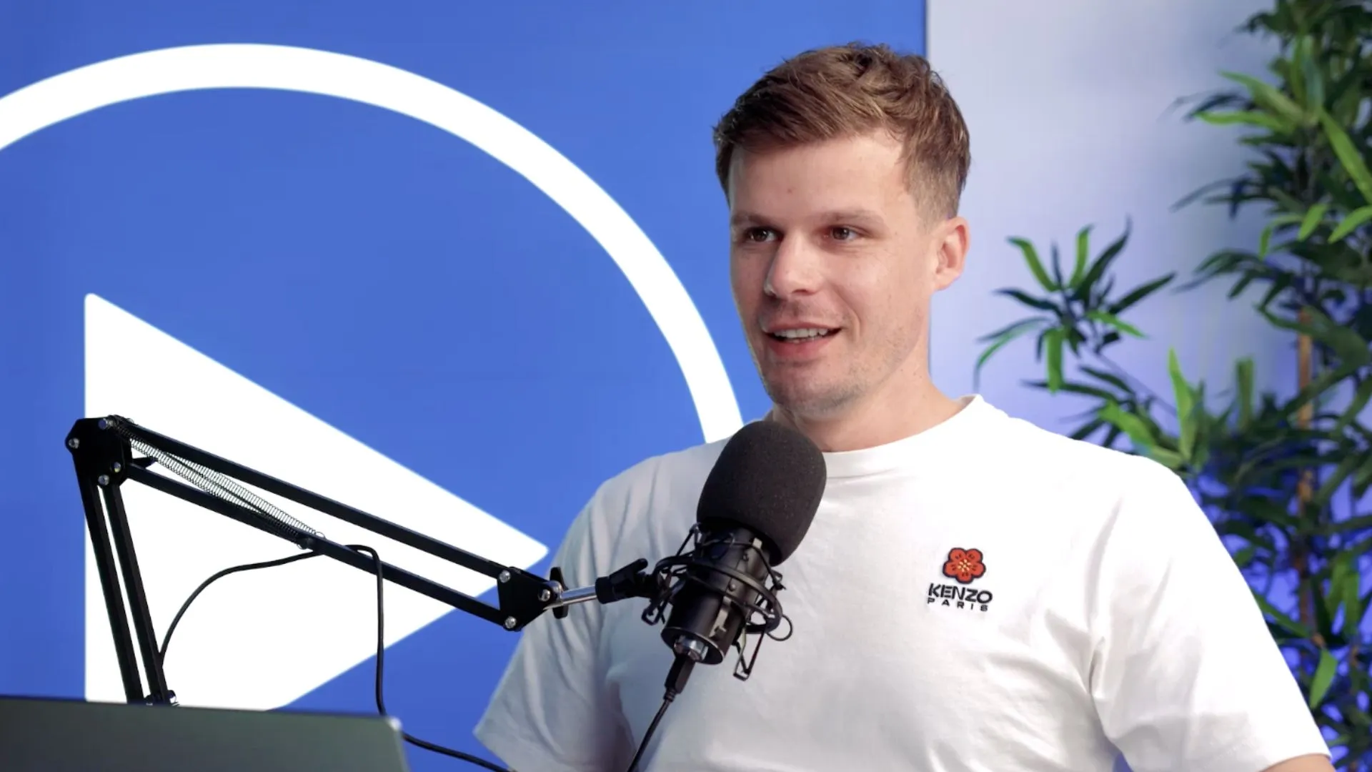 Florian Semmler im Studio mit Mikrofon vor blauer Wand mit Play-Icon