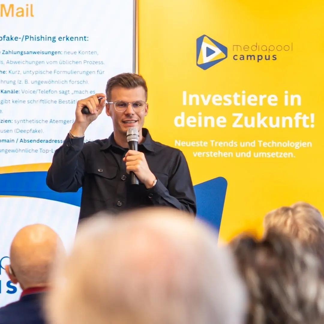 Vortrag zu Phishing und Deepfakes: Referent mit Brille und Mikrofon vor Leinwand und gelbem mediapool campus-Banner, Publikum im Vordergrund