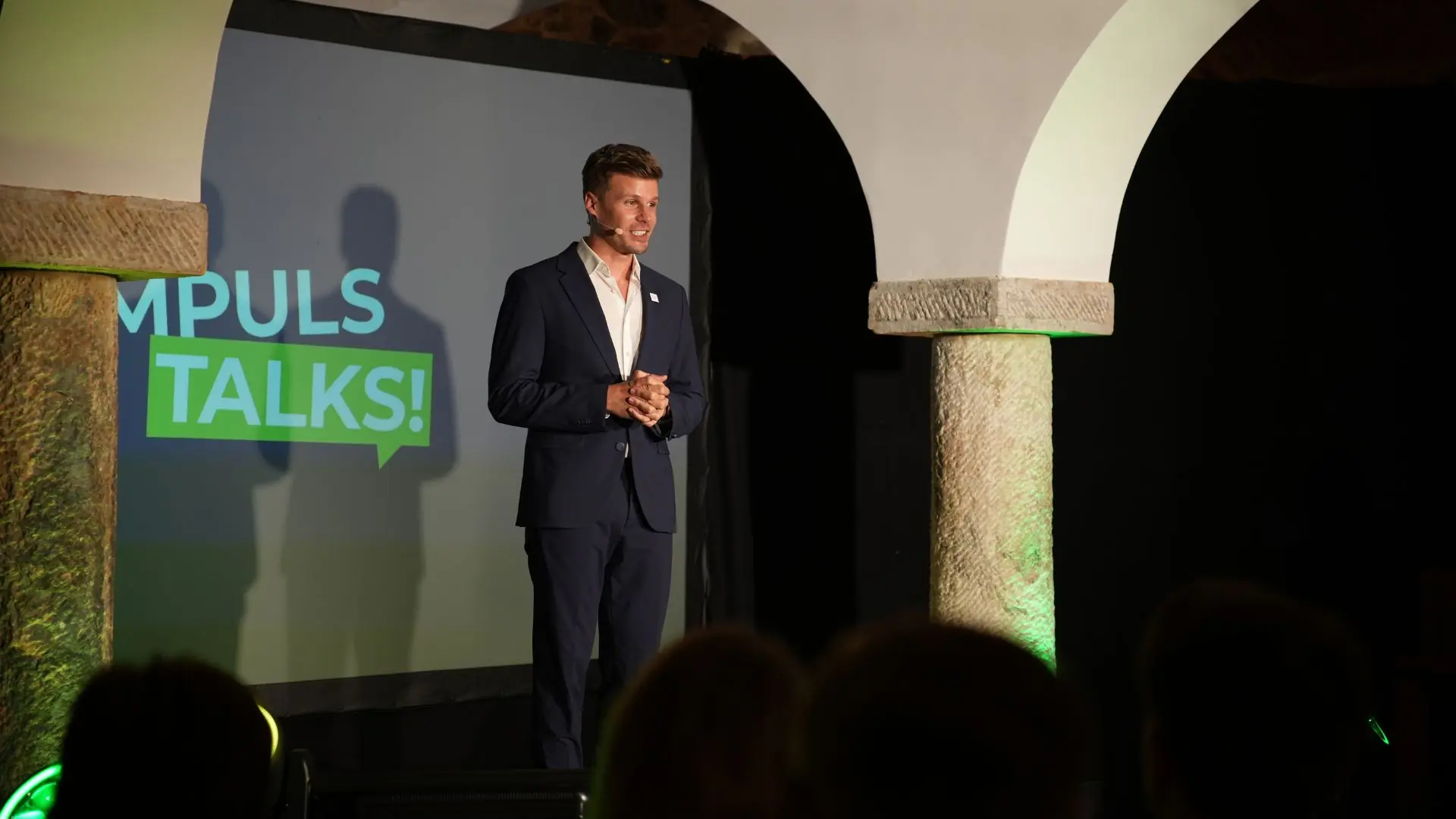 Referent in dunklem Anzug auf der Bühne, Leinwand mit „IMPULS TALKS“, grün angestrahltes Gewölbe
