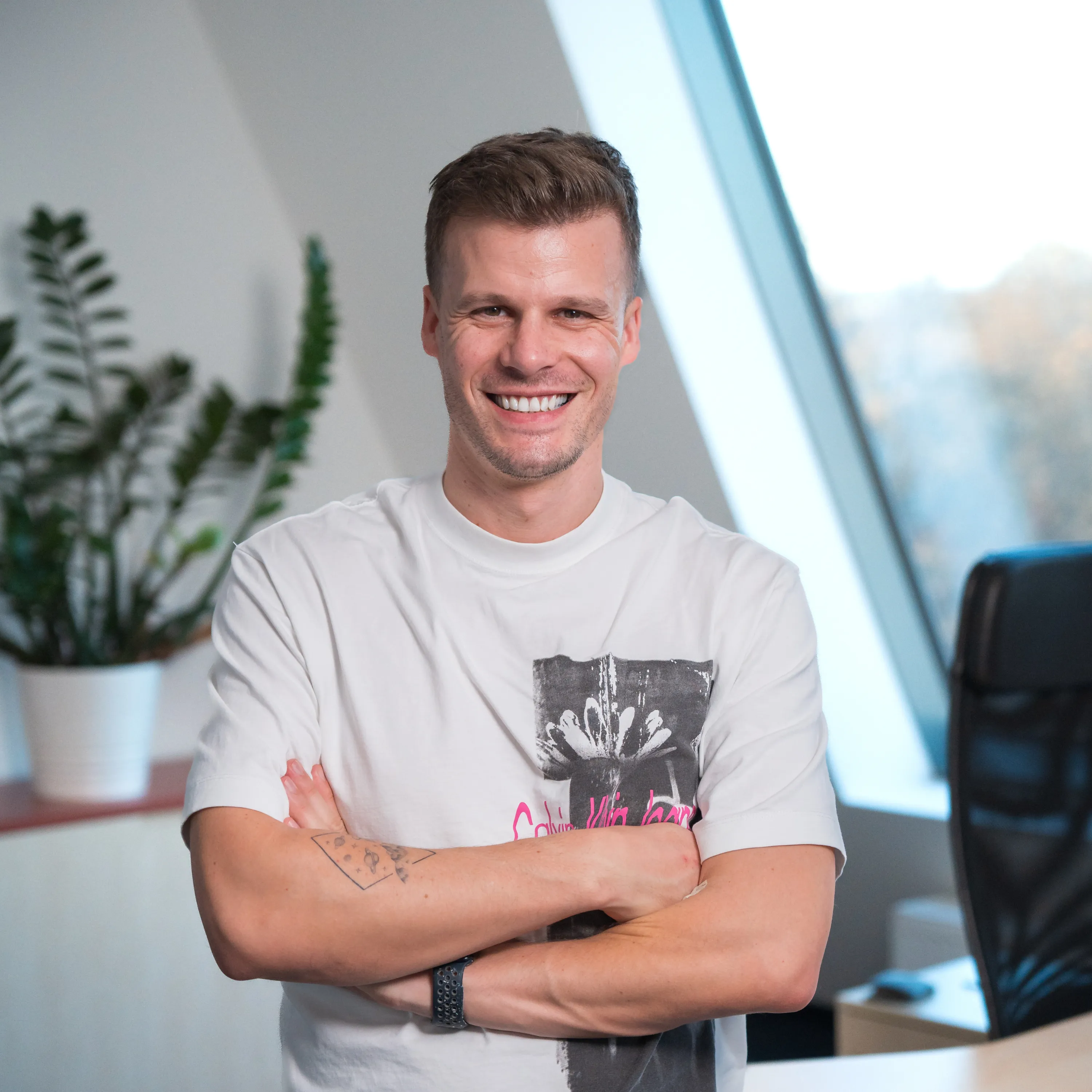 Florian Semmler lächelt mit verschränkten Armen im hellen Büro, Fensterlicht und Zimmerpflanze im Hintergrund